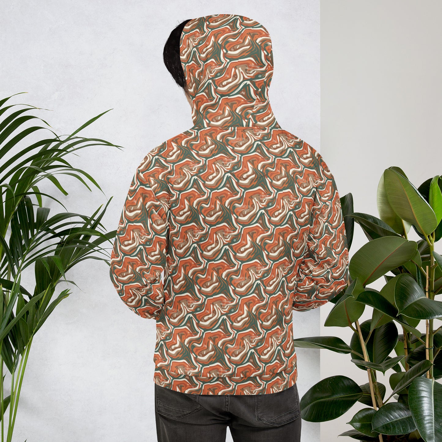 Person wearing a patterned hoodie standing between two plants against a white wall
