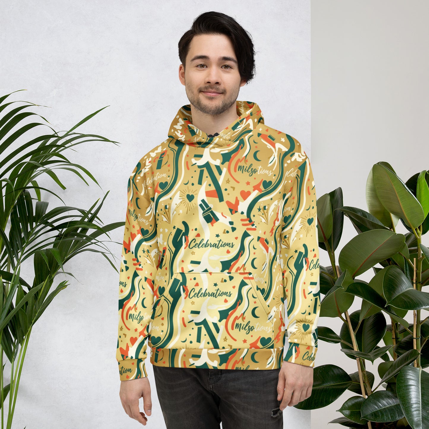Man wearing a colorful hoodie with a pattern, standing between two plants against a white wall.