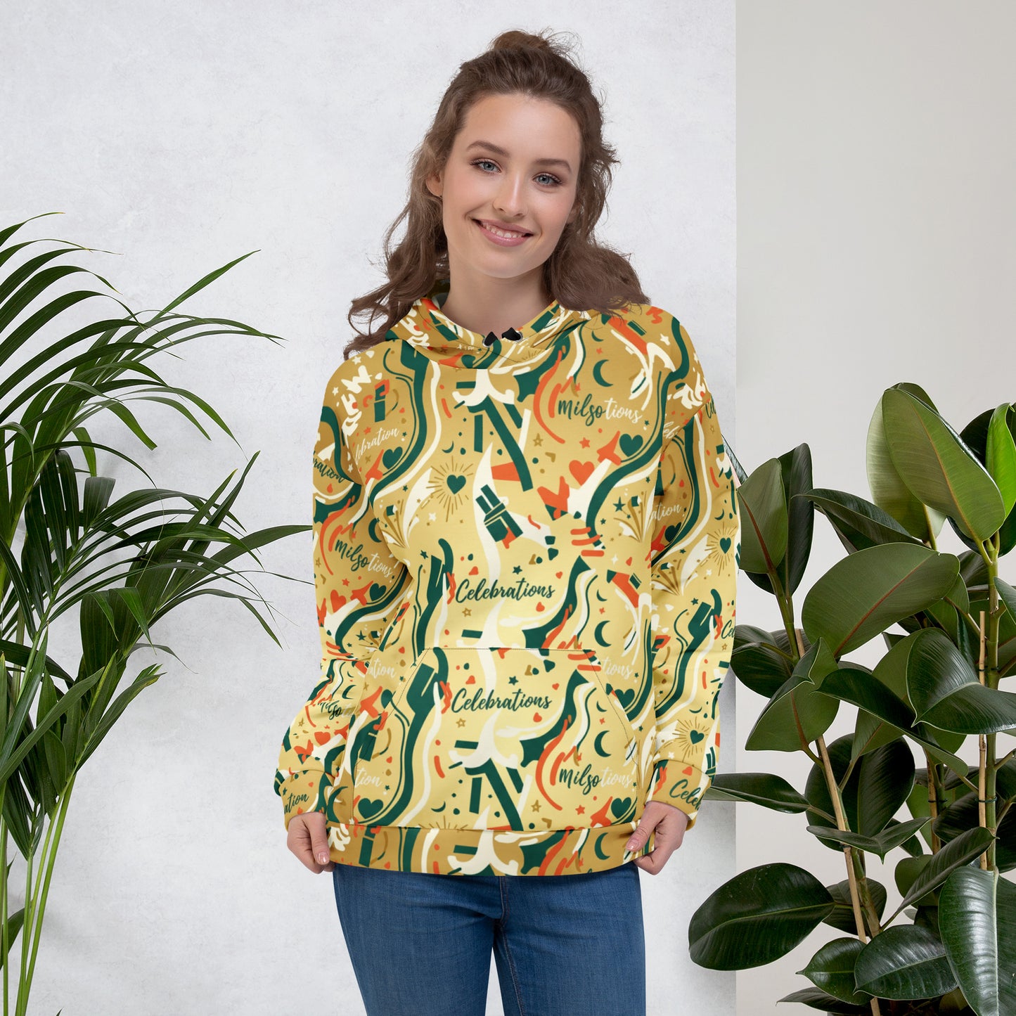 Woman wearing a colorful hoodie with decorative patterns, standing between two plants against a white wall.