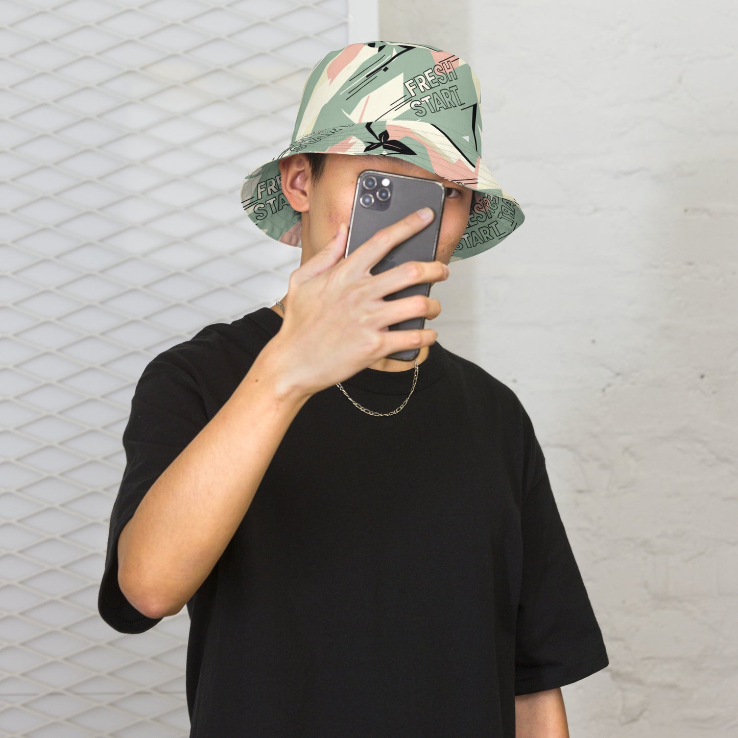 Person wearing a camouflage bucket hat and black t-shirt, taking a selfie against a textured wall.