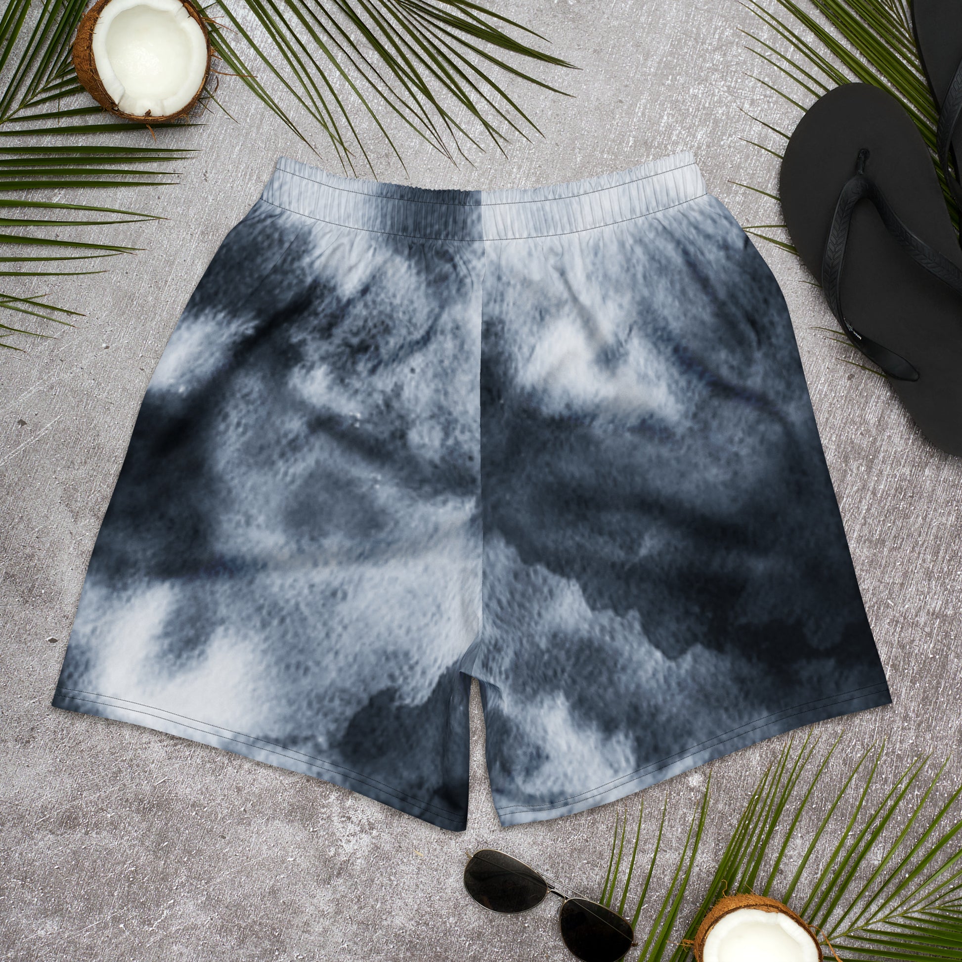 Tie-dye shorts on a textured surface with palm leaves and sunglasses.
