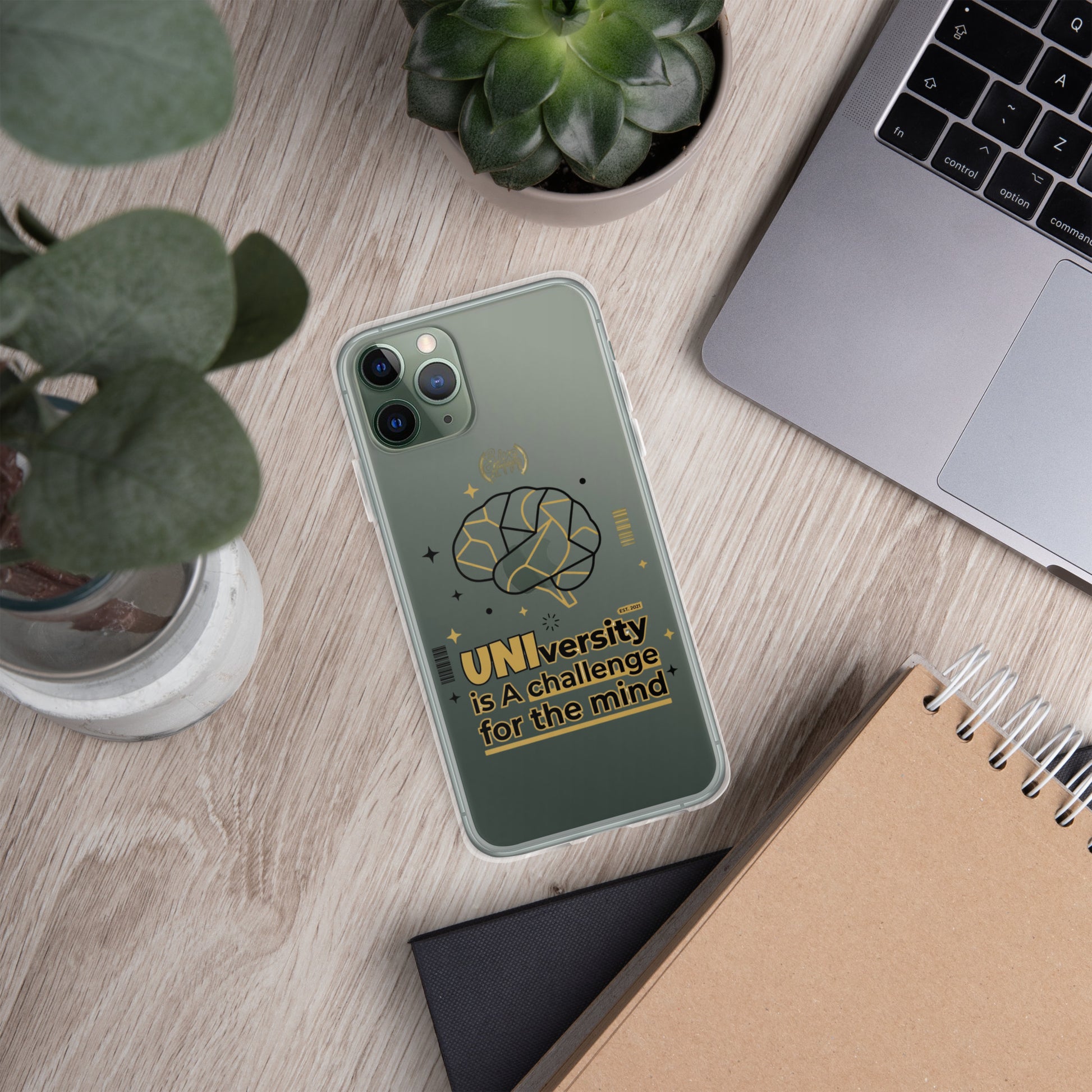 Phone case with Apple logo and 'Make it happen' text on a desk with laptop, notebook, and plants.