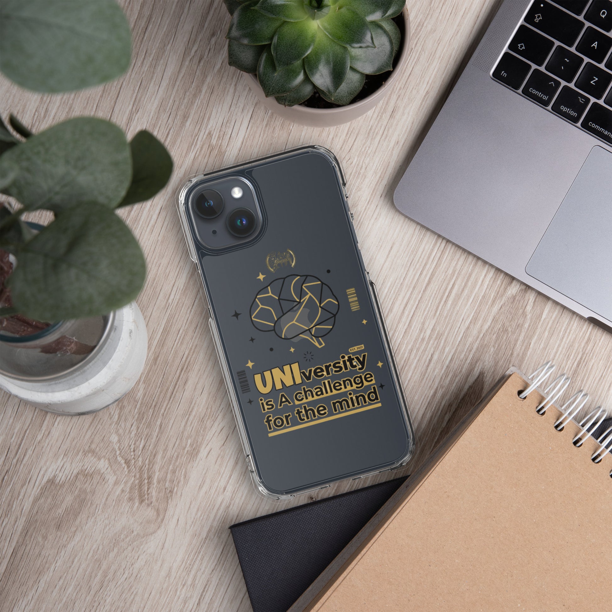 Phone case with 'UNI'versity is a challenge for the mind' text on a desk with laptop, notebook, and plants.