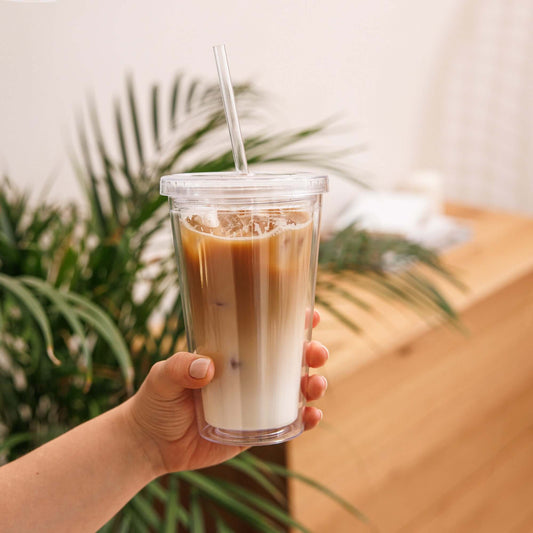 Hand holding a clear plastic cup with iced coffee against a blurred natural background