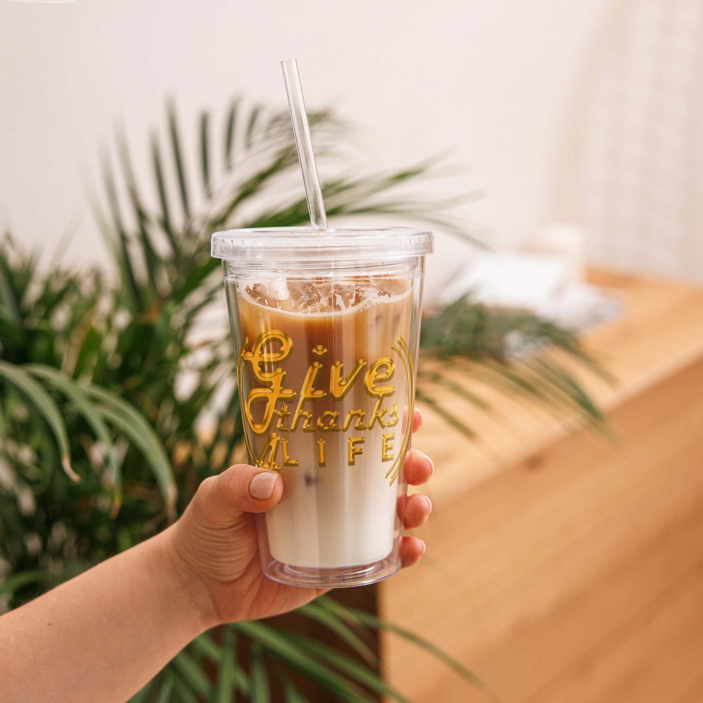 Hand holding a clear cup with iced coffee and 'Give Thanks Life' text, against a blurred indoor background.