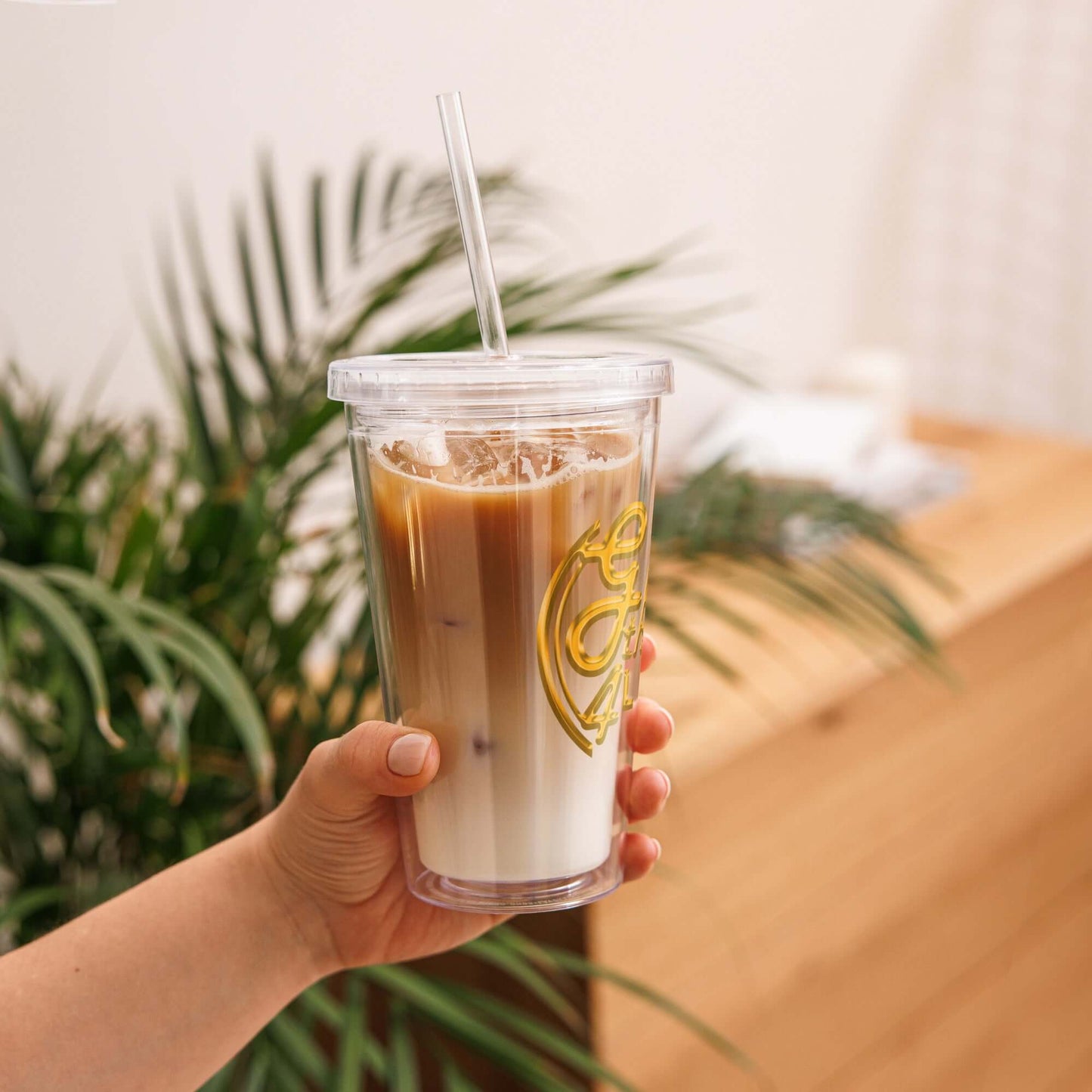 Hand holding a clear cup with iced coffee and a straw, against a blurred natural background.