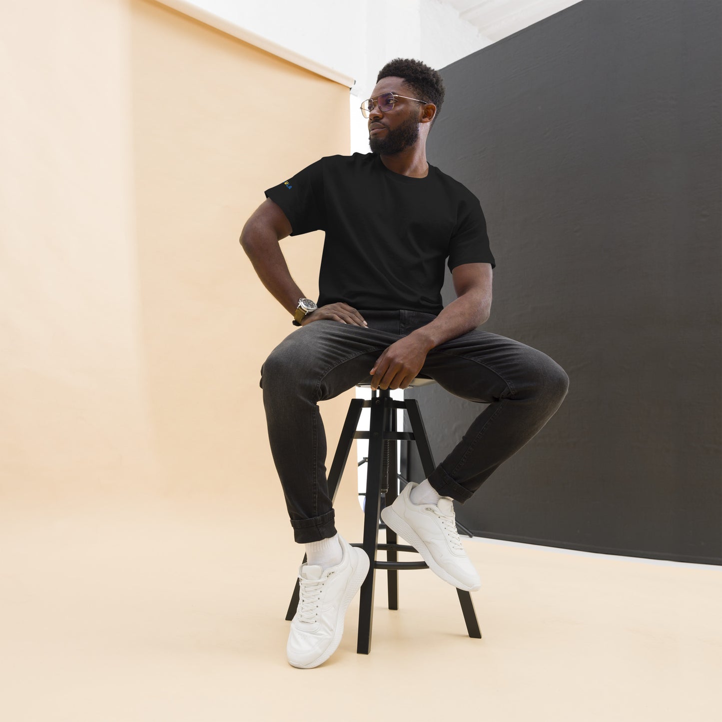 Man sitting on a stool wearing a black t-shirt, dark pants, and white sneakers against a plain background.