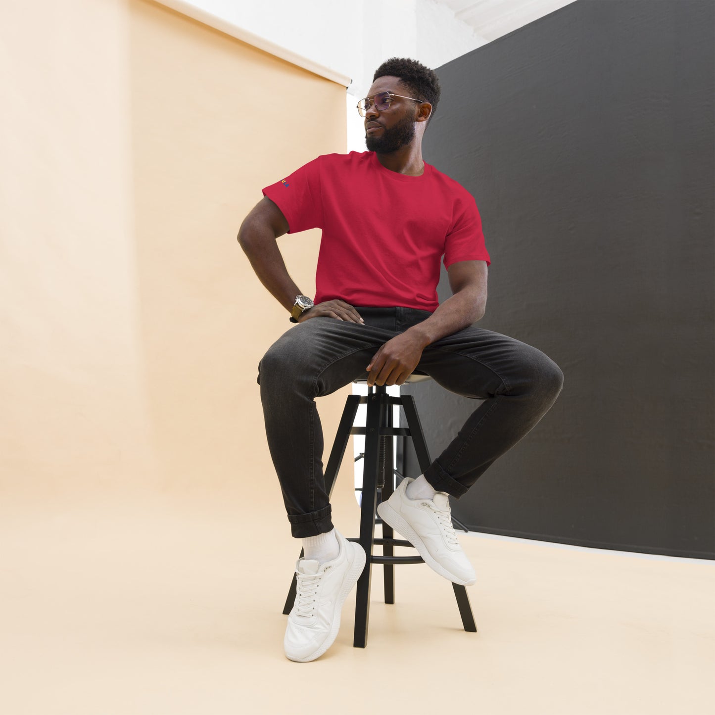 Man sitting on a stool wearing a red shirt, black pants, and white sneakers against a plain background