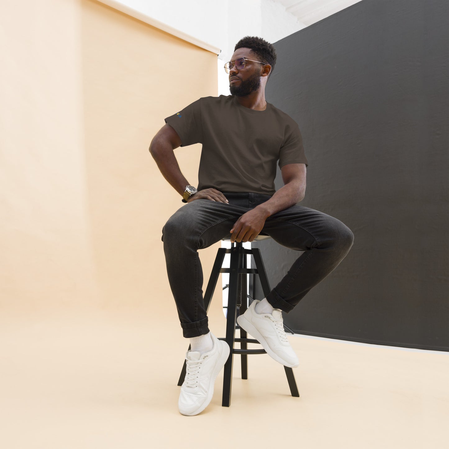 Man sitting on a stool wearing a brown t-shirt, black pants, and white sneakers against a plain background.
