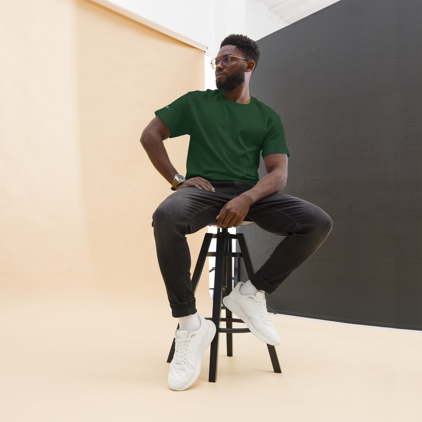 Man sitting on a stool wearing a green t-shirt, black pants, and white sneakers against a plain background.