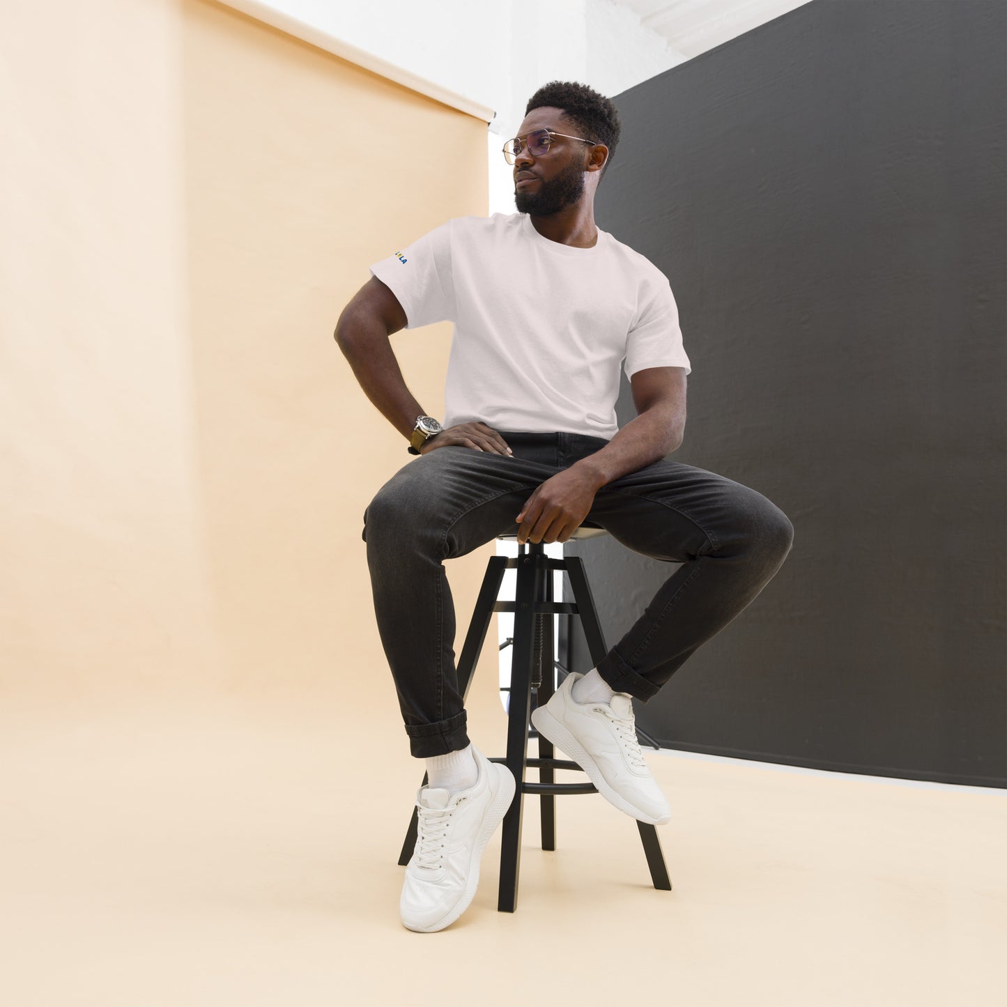 Man sitting on a stool wearing a white t-shirt, black pants, and white sneakers against a plain background