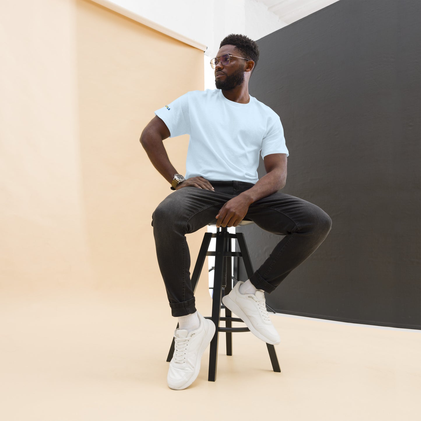 Man sitting on a stool wearing a light blue t-shirt, dark pants, and white sneakers against a plain background.