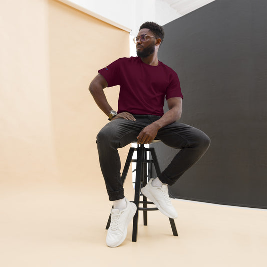 Man sitting on a stool wearing a maroon t-shirt, black pants, and white sneakers against a plain background.