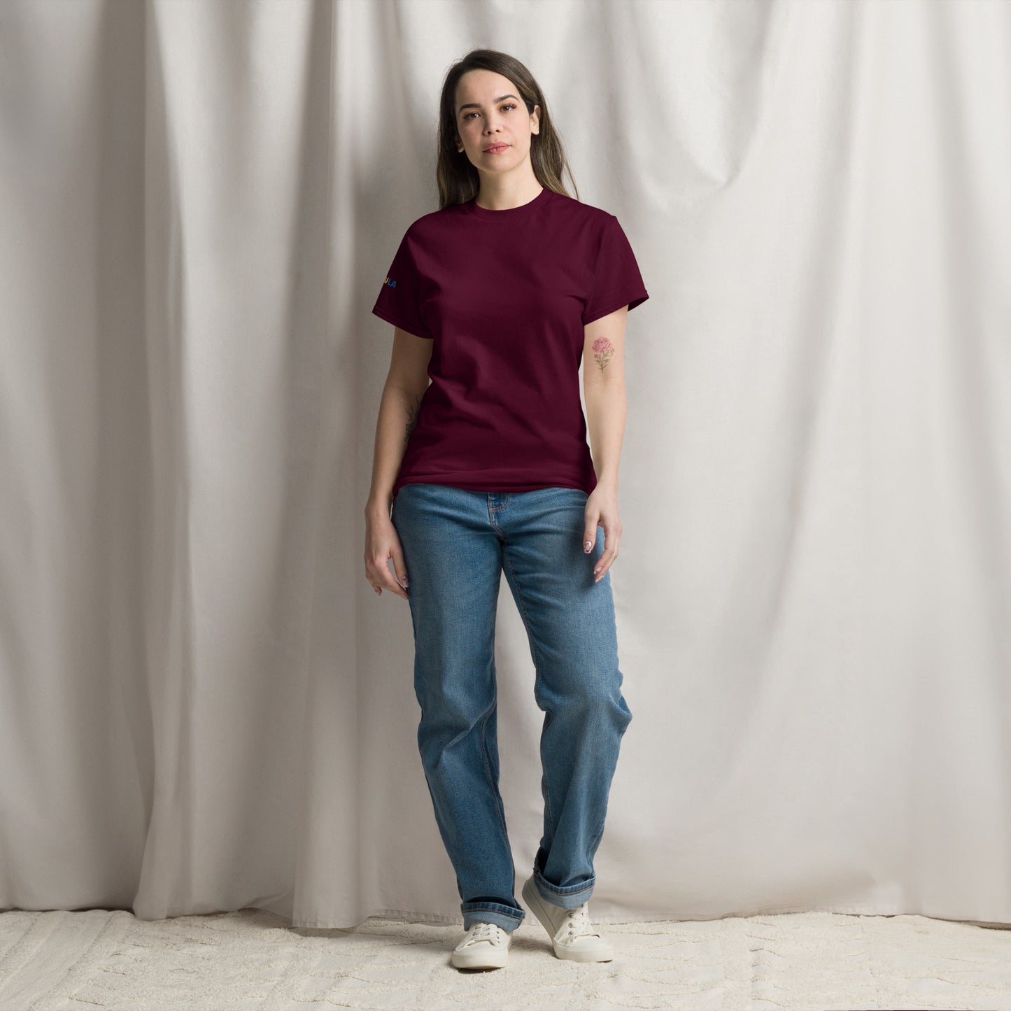 Woman wearing a maroon t-shirt and blue jeans standing against a plain white curtain.