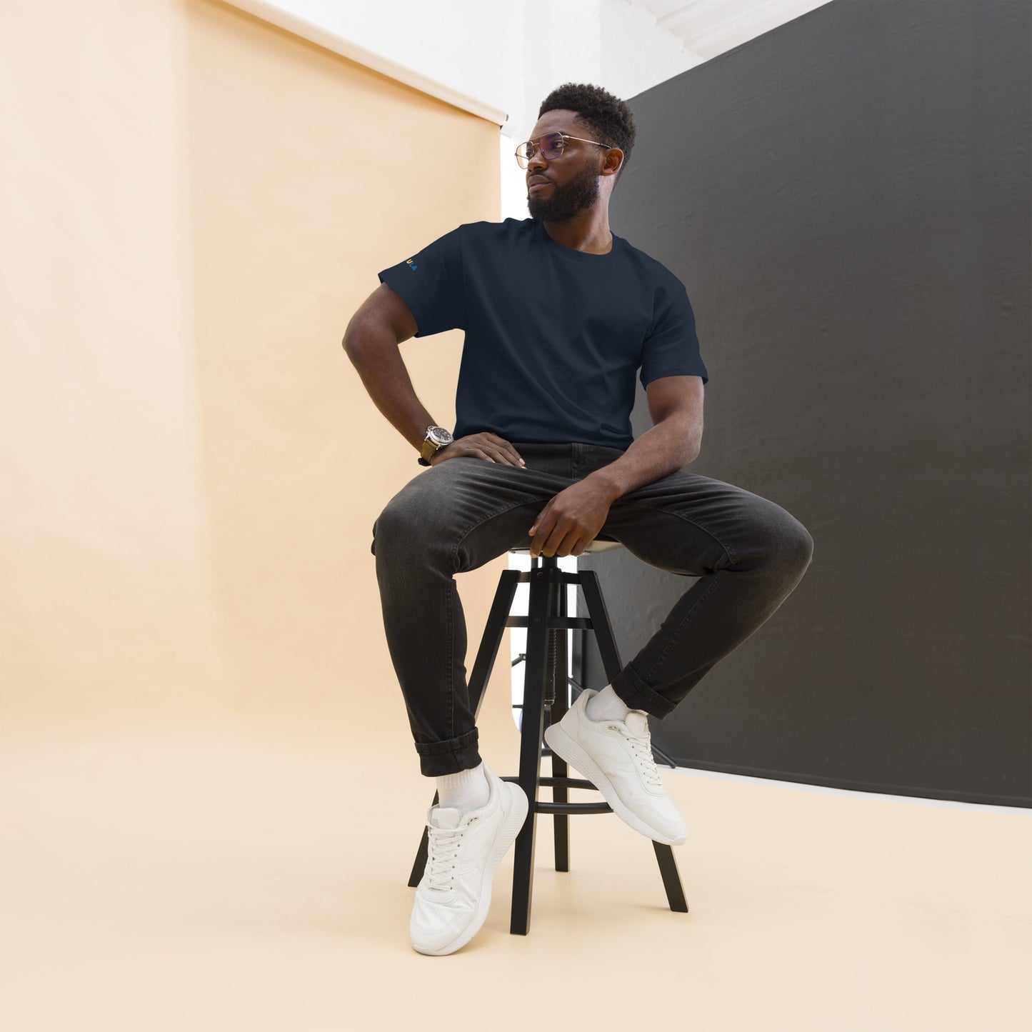Man sitting on a stool wearing a navy t-shirt, dark pants, and white sneakers against a plain background