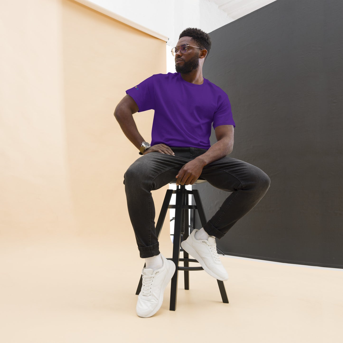 Man sitting on a stool wearing a purple shirt, black pants, and white sneakers against a plain background