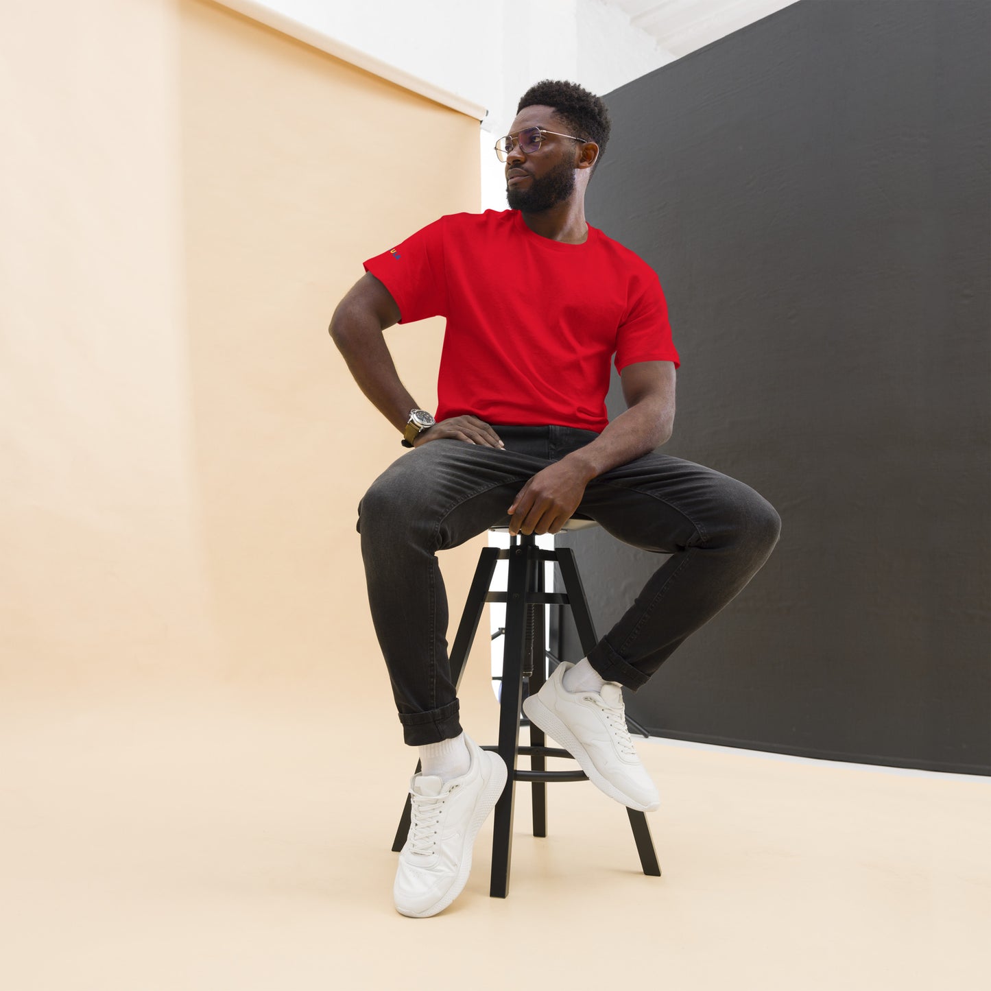 Man sitting on a stool wearing a red shirt, black pants, and white sneakers against a plain background