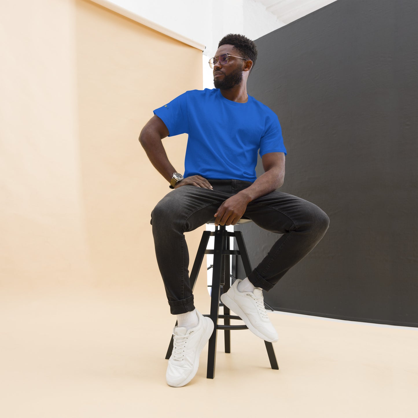 Man sitting on a stool wearing a blue t-shirt, black pants, and white sneakers against a plain background.
