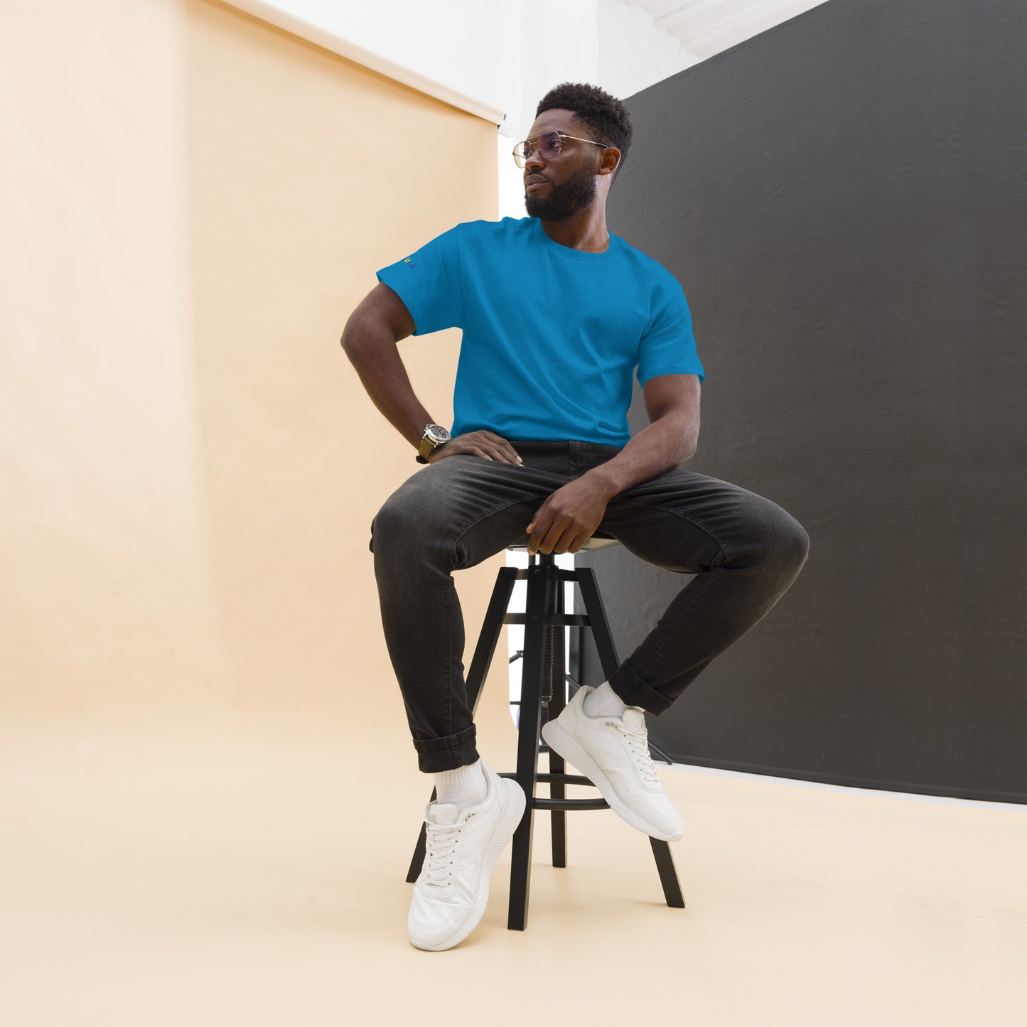 Man sitting on a stool wearing a blue t-shirt, black pants, and white sneakers against a plain background.