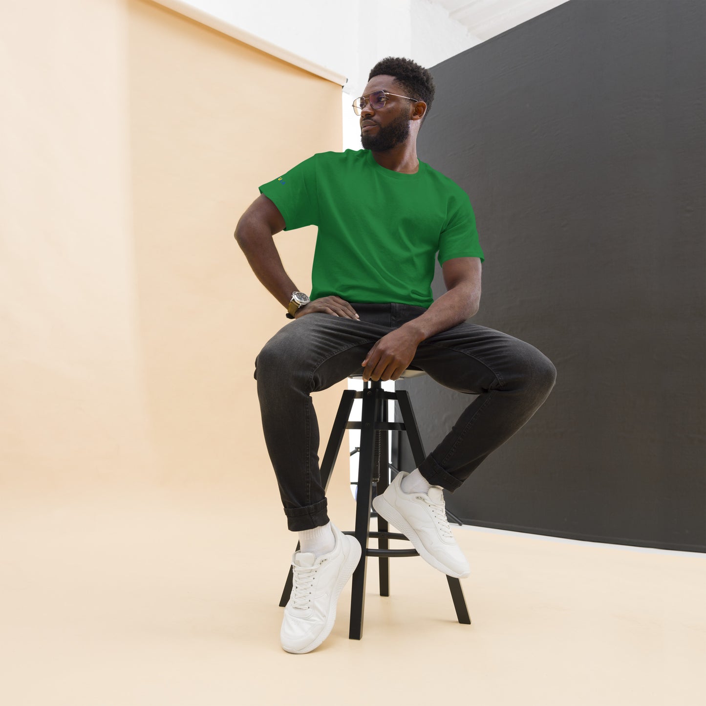 Man sitting on a stool wearing a green t-shirt, black pants, and white sneakers against a plain background.