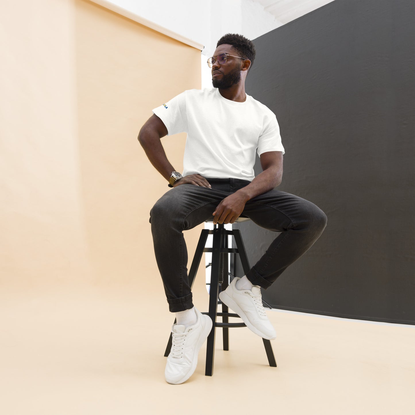 Man sitting on a stool wearing a white t-shirt, black pants, and white sneakers against a plain background