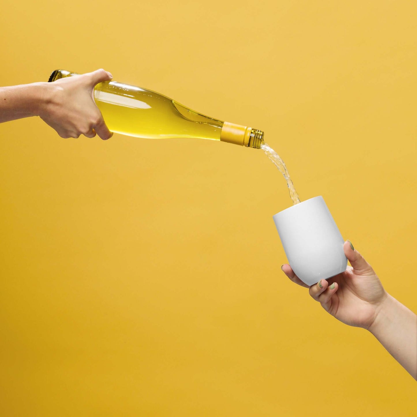 Person pouring white wine from a bottle into a white cup against a yellow background
