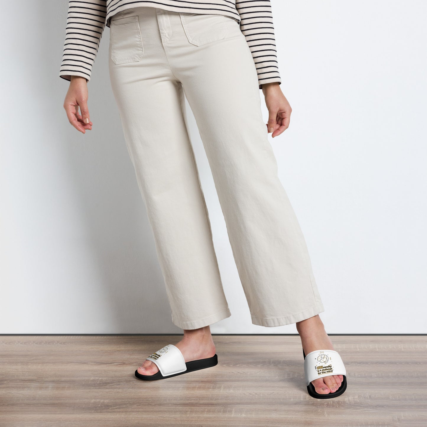 Person wearing white slides with text, beige pants, and a striped shirt on a plain background