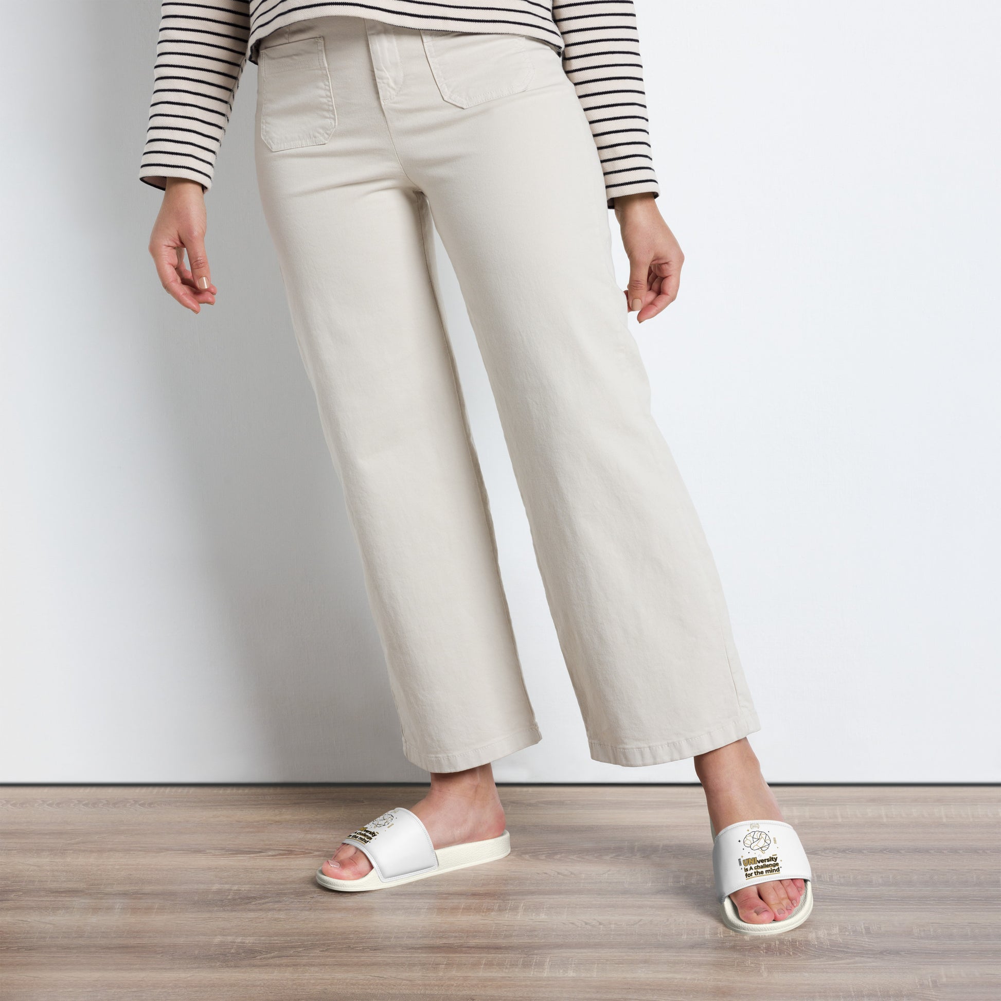 Person wearing white slides, beige pants, and a striped shirt on a wooden floor.