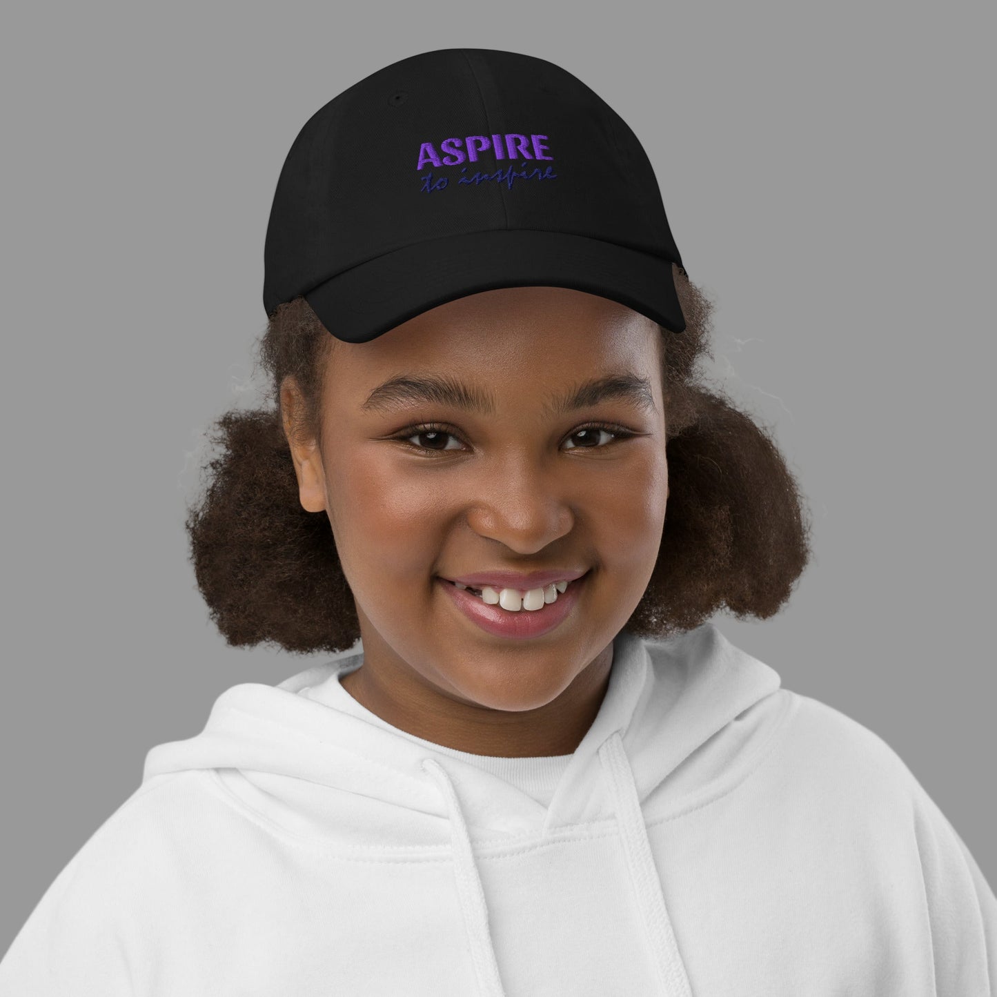 Young girl wearing a black cap with 'ASPIRE' text on a white background