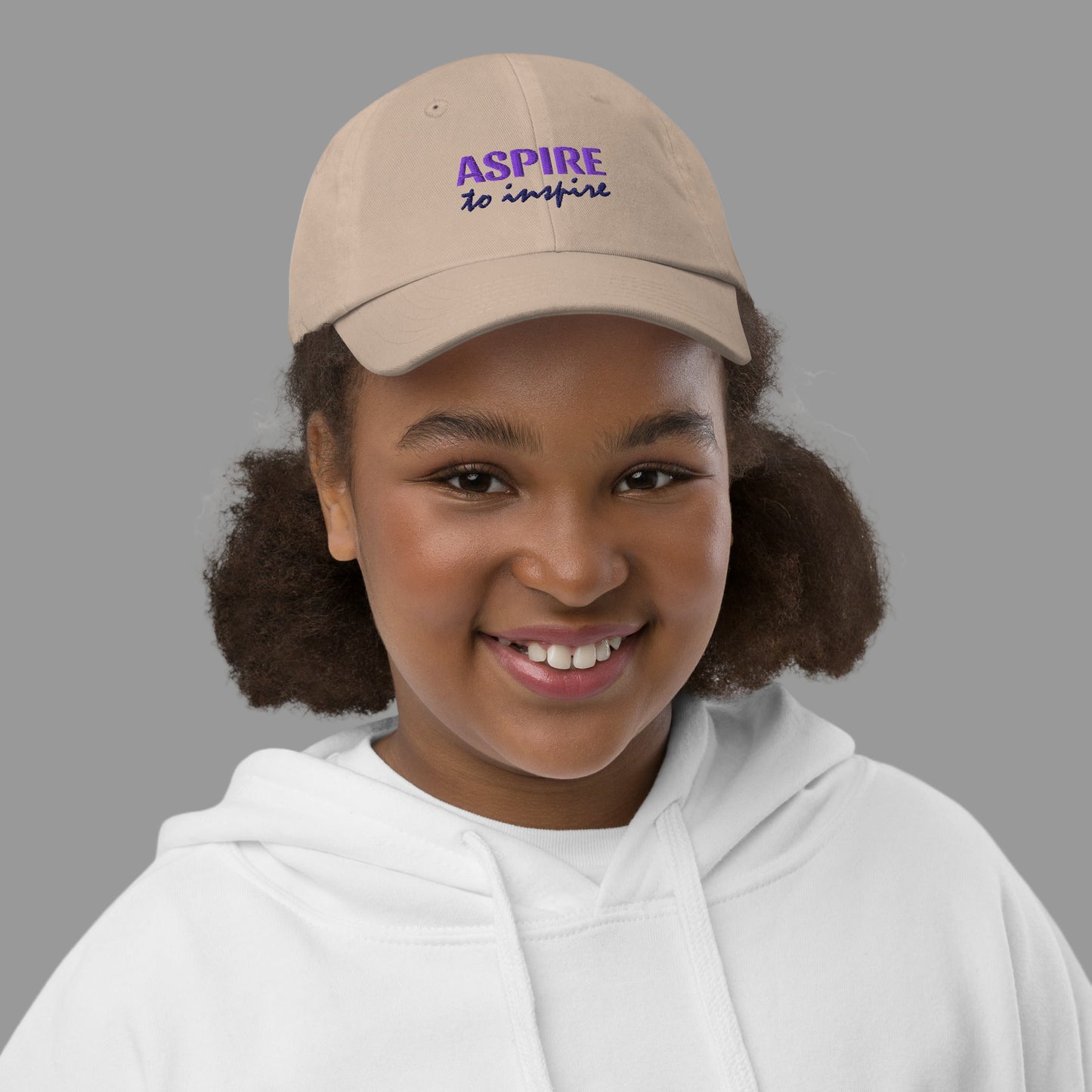 Young girl wearing a beige cap with 'ASPIRE' text on a white background