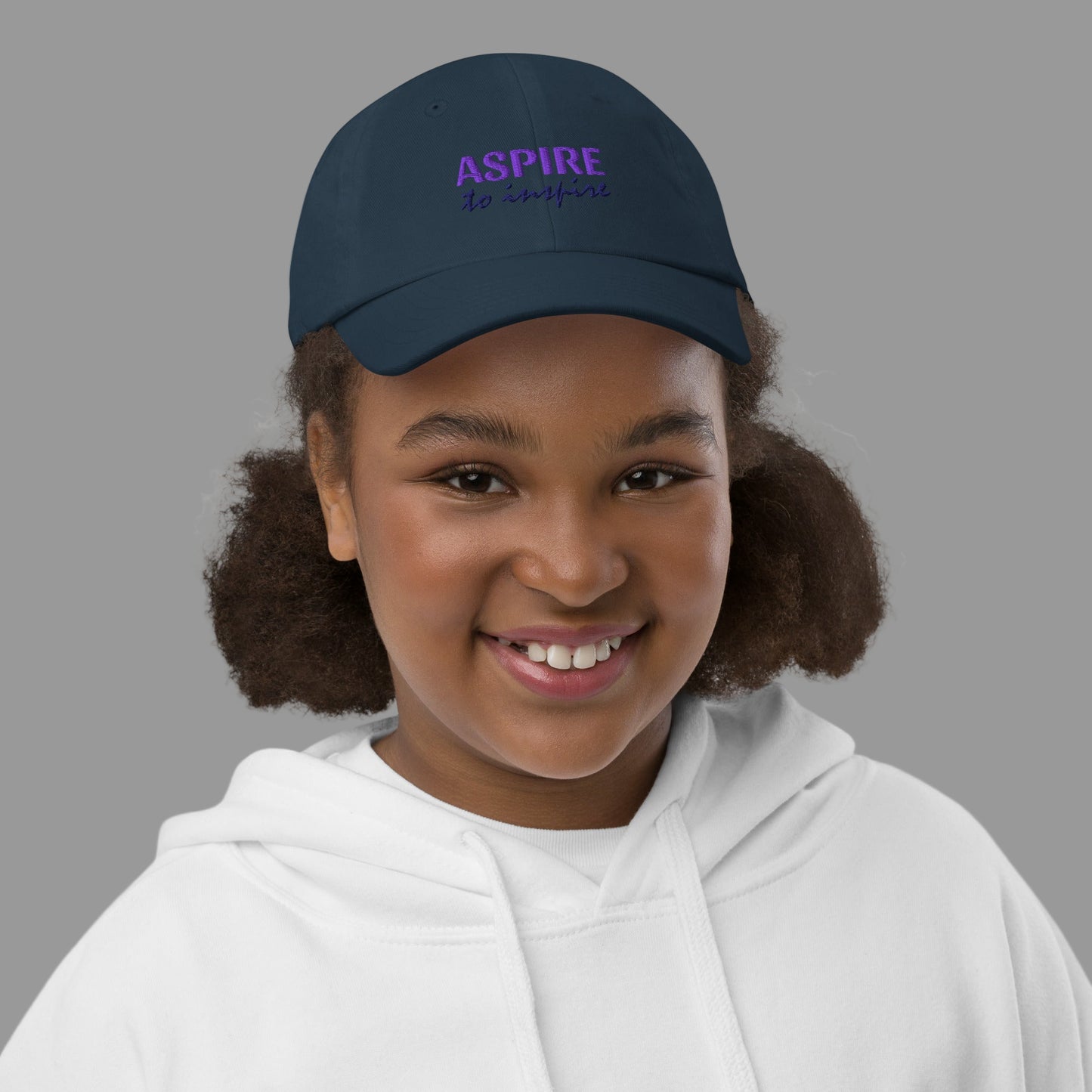 Young girl wearing a navy blue cap with 'ASPIRE' text on a white background