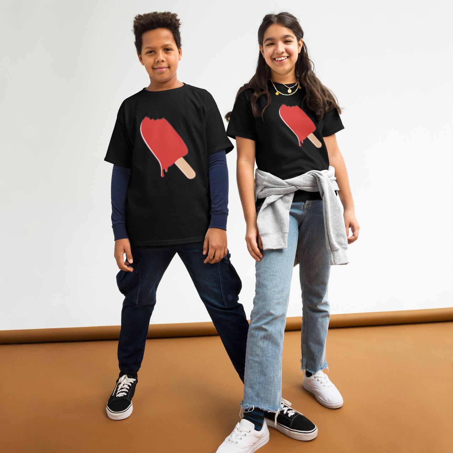 Two children wearing black t-shirts with a red ice cream popsicle design, standing on a brown floor.