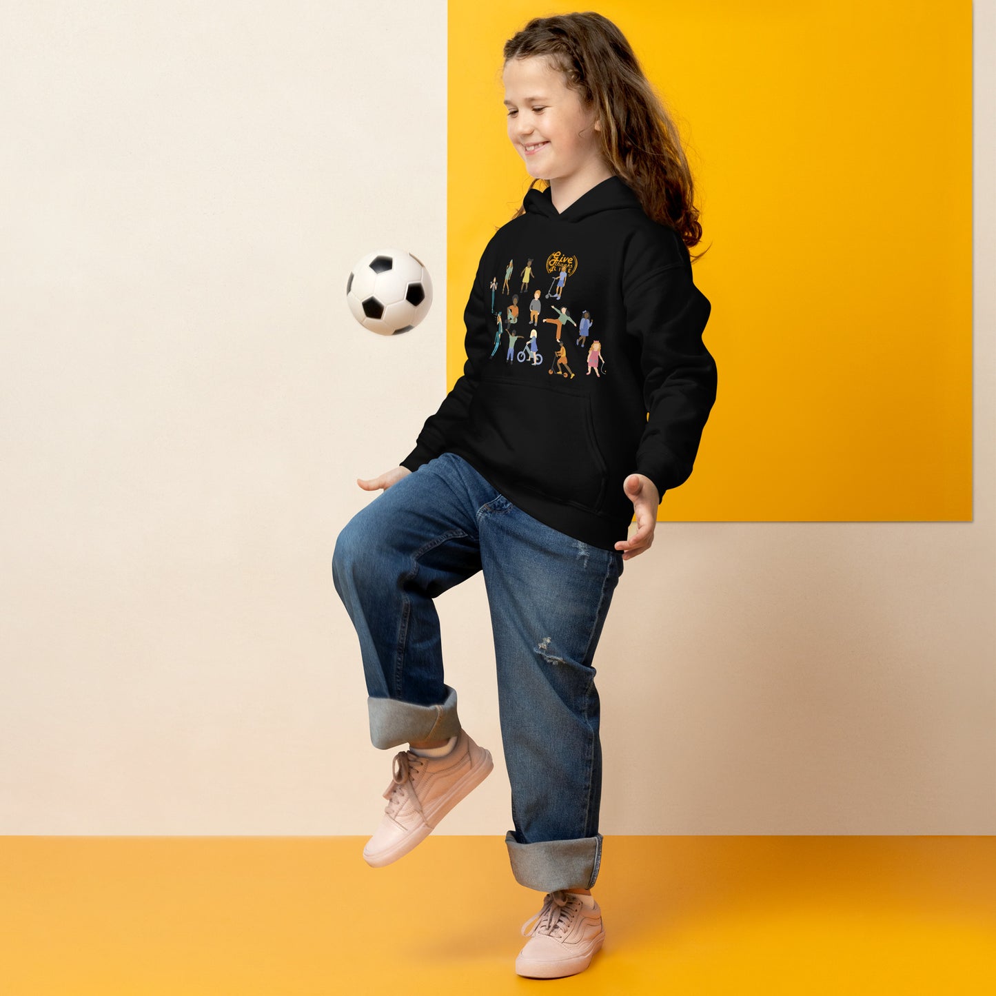 Child wearing a black hoodie with colorful graphics, kicking a soccer ball against a yellow and beige background