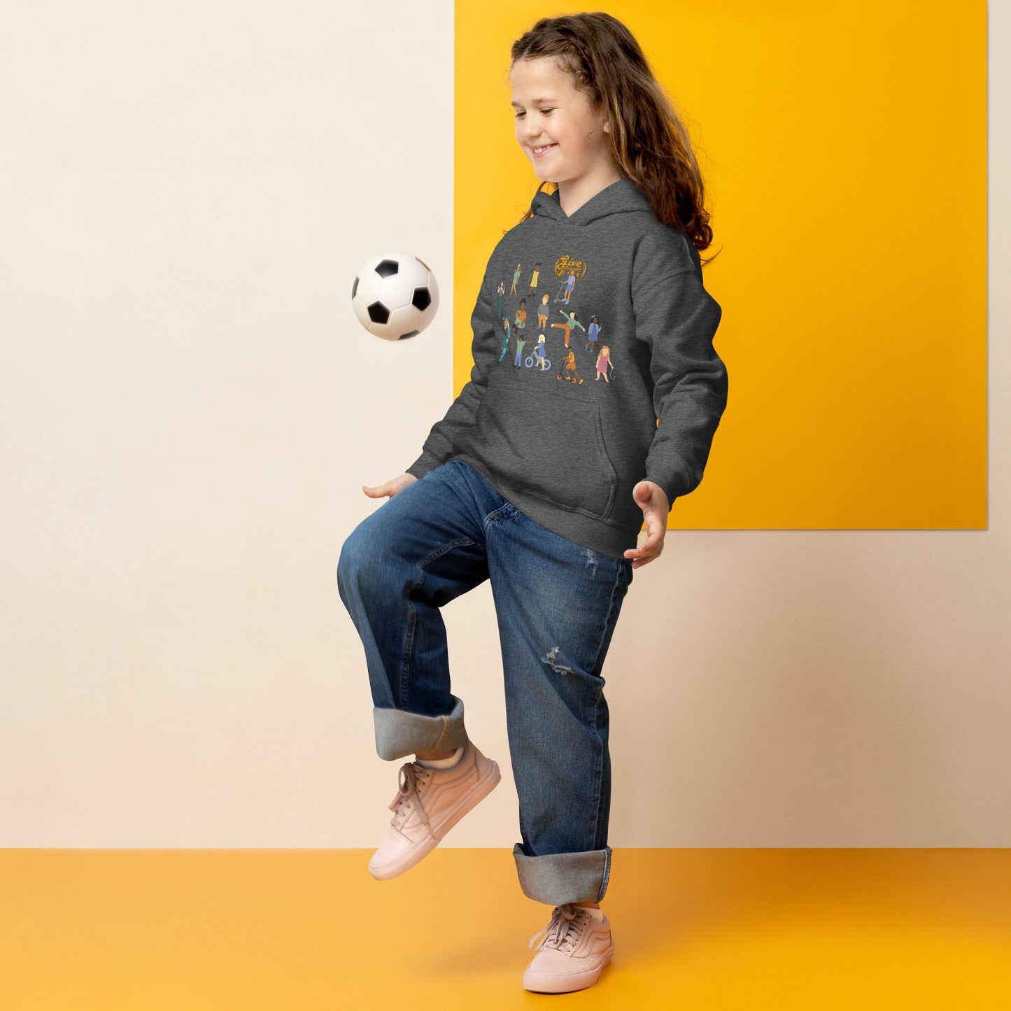 Child wearing a dark gray hoodie with colorful graphics, blue jeans, and white sneakers, standing against a yellow wall.