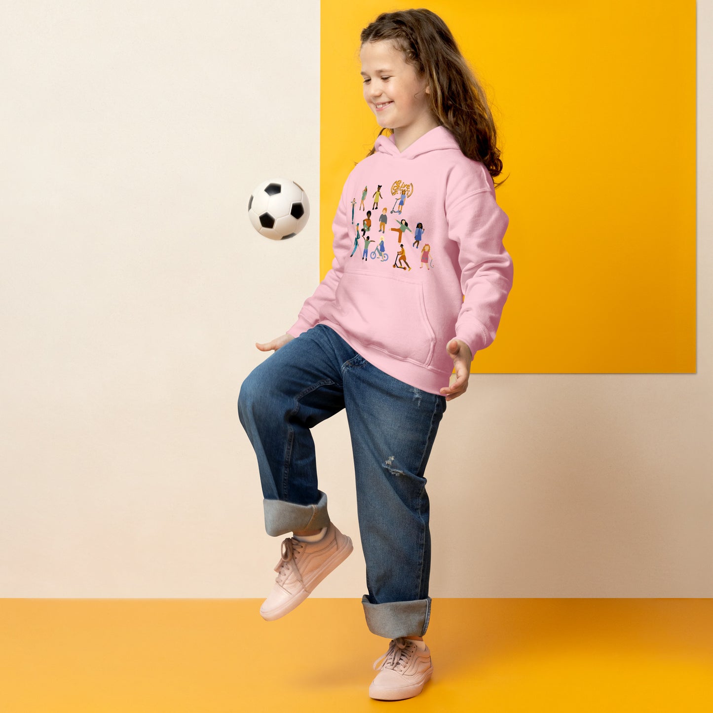 Child wearing a pink hoodie with character design, kicking a soccer ball against a yellow and beige background