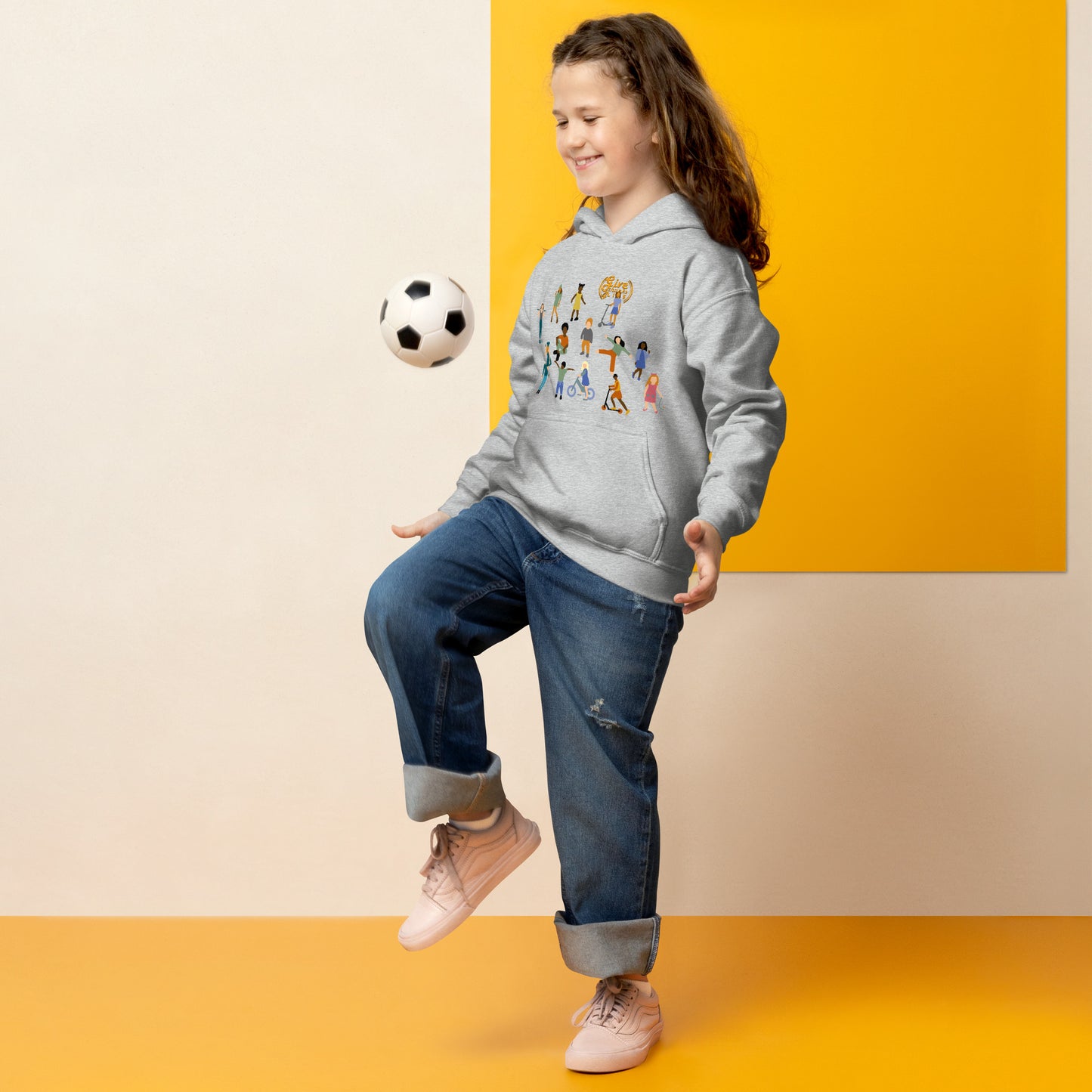Child wearing a gray hoodie with colorful graphics, blue jeans, and pink shoes, kicking a soccer ball against a yellow and beige background.