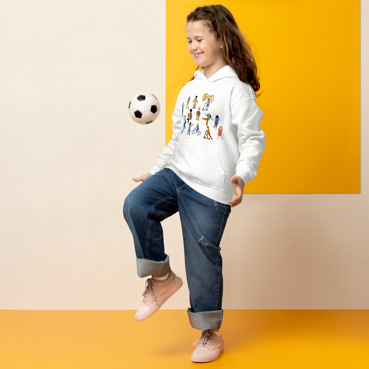 Child wearing a white hoodie with colorful graphics, blue jeans, and pink shoes, kicking a soccer ball against a yellow and beige background.
